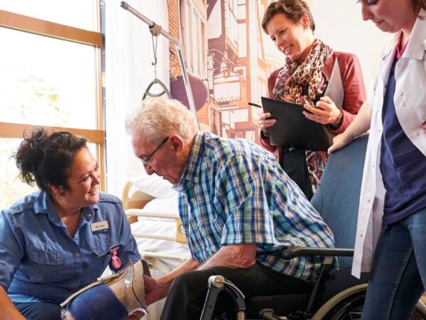 Student oefent zorgactiviteit bij oudere cliënt terwijl docent en begeleider toekijken.